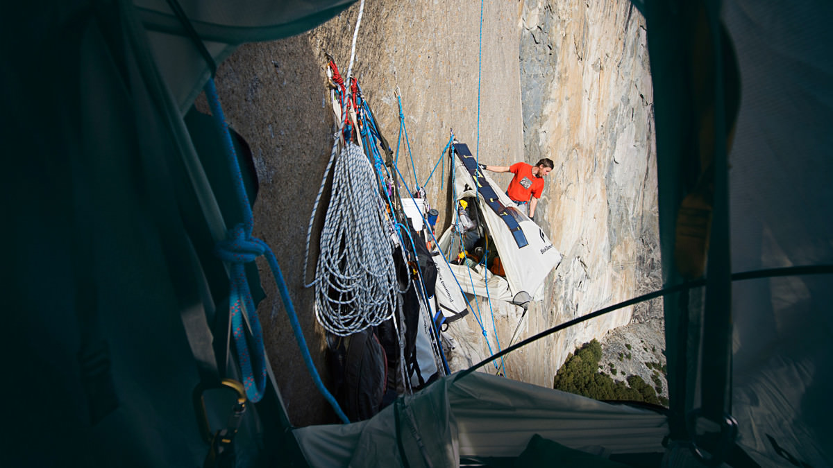 Вид из подвесной платформы. Кевин Йоргесон (Kevin Jorgeson) фотографирует процесс, когда Томми Колдвелл (Tommy Caldwell) занимается позиционированием зарядным устройством "Goal Zero" работающем от солнечных батарей. Эта  зарядка использовалась скалолазами для поддержания питания всех их электронных устройств.  В то время как прямые солнечные лучи на больших стенах вредны для рук (они часто потеют и скользят в зацепах), вредны для скальников (когда размягчается резина и сцепление с зацепом становится неустойчивым. Но в то же время солнце необходимо скалолазам для зарядки электронных устройств. Photo: Corey Rich/Aurora Photos